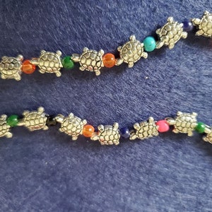 Op de afbeelding: Twee strengen sieraden met zilveren schildpadbedels afgewisseld met kleurrijke kralen. De kralen zijn in verschillende kleuren, waaronder oranje, groen, blauw en roze. De achtergrond is effen donkerblauw.