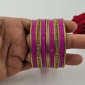 May include: A stack of magenta bangles with gold-colored accents and embedded rhinestones. The bangles are held in a hand against a white background. The bangles are a traditional style of jewelry.
