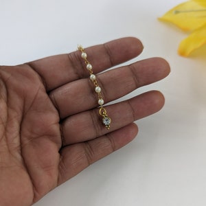 May include: A gold-coloured headpiece with a string of small, white pearls. The strand ends in a teardrop-shaped pendant with a clear, sparkling stone. The jewellery is designed for the forehead, possibly for a traditional or cultural outfit.