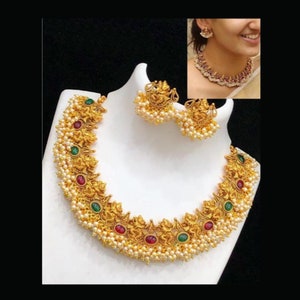 May include: A gold-toned necklace and matching earrings set displayed on a white bust. The necklace features intricate detailing, red and green gemstones, and small white pearls. The earrings have a similar design. A smaller image shows the necklace being worn.