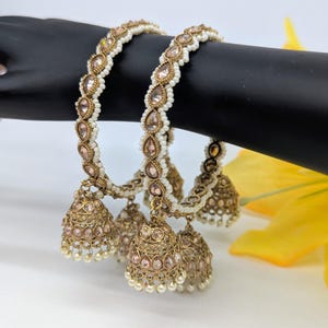 May include: Two ornate gold bangles with dangling bell charms. The bangles are adorned with white beads and teardrop-shaped pink gemstones. The bell charms are detailed with pink stones and small white pearls.