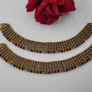 Op de afbeelding: Twee goudkleurige enkelbandjes met ingewikkelde details en kleurrijke kralen. De enkelbandjes hebben een kettingontwerp met rijen kleine, ronde versieringen en een franje van veelkleurige kralen. Een rode roos staat op de achtergrond.