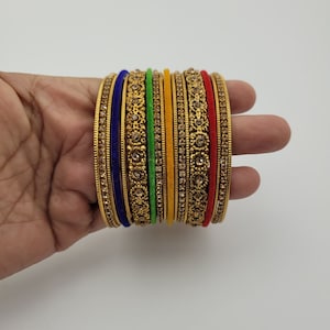 Puede incluir: Un conjunto de pulseras doradas con intrincados dise&ntilde;os florales y gemas incrustadas. El conjunto incluye pulseras en varios colores, como azul, verde, amarillo y rojo, creando un aspecto vibrante y ornamentado. Las pulseras se sostienen en una mano.