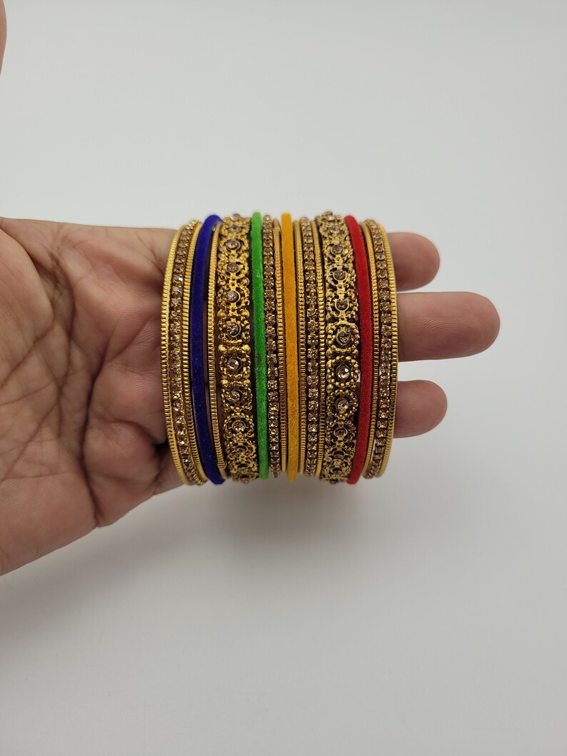 Puede incluir: Un conjunto de pulseras doradas con intrincados dise&ntilde;os florales y gemas incrustadas. El conjunto incluye pulseras en varios colores, como azul, verde, amarillo y rojo, creando un aspecto vibrante y ornamentado. Las pulseras se sostienen en una mano.