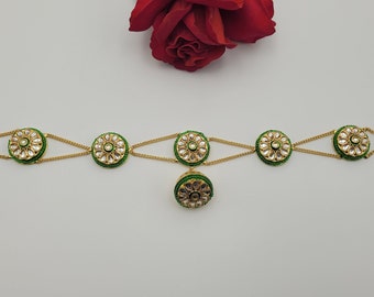 May include: A gold-coloured headpiece with green and white detailing. The design incorporates circular elements with intricate patterns, linked by fine chains. A central pendant hangs down. A red rose is in the background.