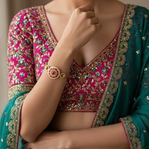 May include: A vibrant fuchsia blouse and skirt set featuring intricate gold embroidery and green sequin accents. The ensemble includes a deep teal dupatta with a wide gold embroidered border. A circular gold bracelet with a central red stone is worn.