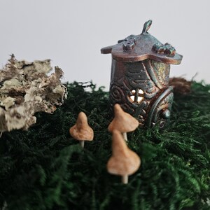 May include: A miniature fairy house made of clay with a green and brown glaze, surrounded by three small brown mushrooms on a bed of green moss.