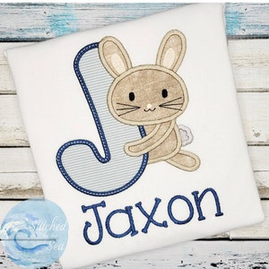 May include: A white onesie with a blue letter J and a brown bunny holding onto the letter. The name Jaxon is embroidered in blue below the letter.