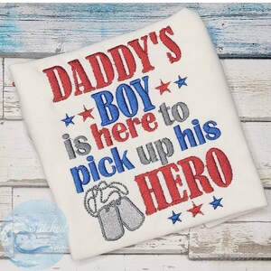 Puede incluir: Camiseta blanca con bordado rojo, azul y gris que dice "Daddy's Boy is here to pick up his HERO" con dos placas de identificaci&oacute;n plateadas y estrellas.