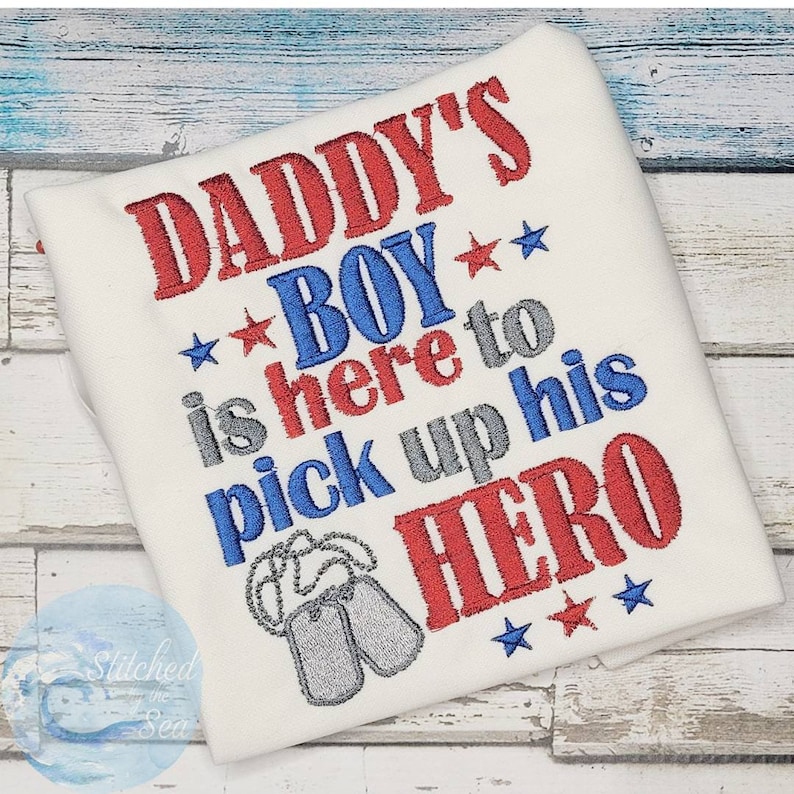Puede incluir: Camiseta blanca con bordado rojo, azul y gris que dice "Daddy's Boy is here to pick up his HERO" con dos placas de identificaci&oacute;n plateadas y estrellas.