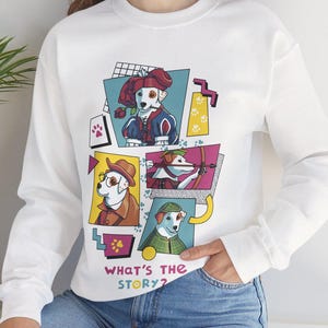 May include: White sweatshirt featuring a colorful graphic of illustrated dogs in various costumes. The design includes the text "WHAT'S THE STORY?" in a playful font. The sweatshirt has long sleeves and a crew neck.