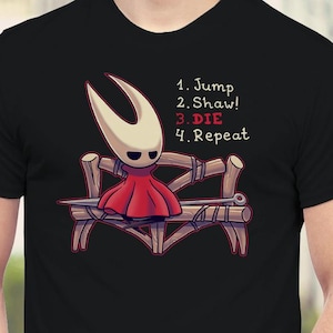 May include: Black t-shirt featuring a cartoon character in a red dress and a white mask, sitting on a wooden bench. The text on the shirt reads: "1. Jump 2. Shaw! 3. Die 4. Repeat."