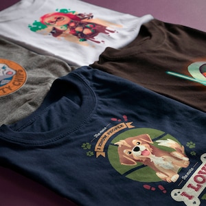 Op de afbeelding: Vier t-shirts in verschillende kleuren, elk met een uniek grafisch ontwerp. Ontwerpen omvatten een stripfiguur, een hond met de tekst "I Love Dogs" en een wezen met een lichtzwaard. De shirts worden gepresenteerd op een paars oppervlak.