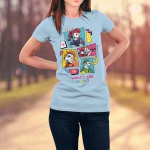 Op de afbeelding: Lichtblauw T-shirt met een grafisch ontwerp van cartoonhonden in verschillende kostuums. Het ontwerp bevat de tekst "WHAT'S THE STORY?" in roze. Het shirt is gemaakt van een zacht materiaal.