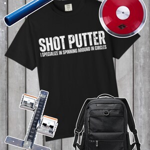 以下が含まれることがあります： 「SHOT PUTTER I SPECIALIZE IN SPINNING AROUND IN CIRCLES」の文字が入った黒いTシャツ。赤い円盤、黒いバックパック、青いバトン、陸上競技のスタートブロックも写っています。