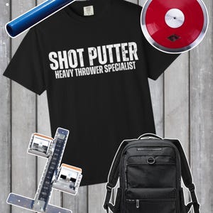 以下が含まれることがあります： 「SHOT PUTTER HEAVY THROWER SPECIALIST」の白文字が入った黒いTシャツ。青いやり、赤い円盤、黒いバックパック、陸上競技のスタートブロックも表示されています。