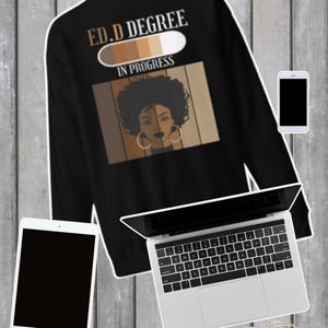 Op de afbeelding: Zwart sweatshirt met de tekst "ED.D DEGREE IN PROGRESS" boven een afbeelding van een vrouwengezicht met bruine huidtinten. Een tablet, een laptop, een telefoon en een bril staan ook op de afbeelding.