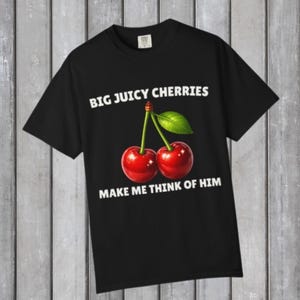 Pode incluir: Uma t-shirt preta de manga curta com um gráfico de duas cerejas vermelho-brilhante com uma folha verde nos talos. O texto "BIG JUICY CHERRIES" está impresso a branco acima e "MAKE ME THINK OF HIM" está impresso a branco abaixo.