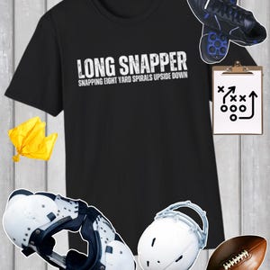 May include: A black t-shirt with white text that reads LONG SNAPPER SNAPPING EIGHT YARD SPIRALS UPSIDE DOWN, surrounded by a yellow penalty flag, black football cleats, a clipboard with a play diagram, white shoulder pads, a white helmet, and a football.