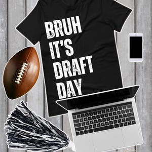 Könnte beinhalten: Schwarzes T-Shirt mit dem weißen Text "BRUH IT'S DRAFT DAY". Ein brauner Football, ein schwarz-weißer Pompon, ein Laptop, ein Smartphone und eine Brille sind ebenfalls im Bild. Die Gegenstände liegen auf einer grauen Holzoberfläche.