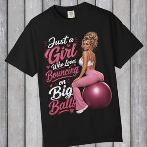 May include: A black t-shirt featuring a graphic of a person with blonde hair in a bun, wearing a white sports bra and pink leggings, sitting on a large purple exercise ball. The text reads: Just a Girl Who Loves Bouncing on Big Balls.