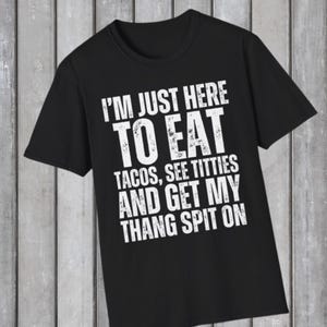 Puede incluir: Camiseta negra con texto blanco: "I'M JUST HERE TO EAT TACOS, SEE TITTIES AND GET MY THANG SPIT ON". La camiseta tiene cuello redondo y mangas cortas. El fondo es una superficie de madera gris claro.