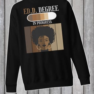 Op de afbeelding: Zwart sweatshirt met de tekst "E.D.D. DEGREE IN PROGRESS" boven een afbeelding van een vrouwengezicht. De afbeelding bevat een kleurenpalet en een gespleten gezichtsontwerp. De vrouw draagt grote oorringen.