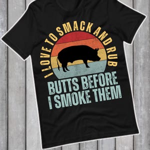 Peut inclure: T-shirt noir avec un motif graphique représentant une silhouette de cochon à l'intérieur d'un cercle rétro au coucher du soleil. Le texte dit "I LOVE TO SMACK AND RUB BUTTS BEFORE I SMOKE THEM."