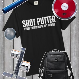 以下が含まれることがあります： 「SHOT PUTTER I LOVE THROWING HEAVY THINGS」の文字が入った黒いTシャツは、スポーツ用品に囲まれています。青いバトン、赤い円盤、黒いバックパック、ショットプットボール、スタートブロックなどがあります。