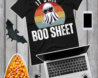 以下が含まれることがあります： ハロウィーンをテーマにしたグラフィックの黒いTシャツ。デザインは、サングラスをかけた幽霊が夕焼け色の円の前にいて、「IT'S ALL BOO SHEET」というテキストが書かれています。追加のハロウィーンの装飾には、キャンディーコーン、クモ、コウモリが含まれています。