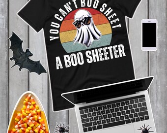 以下が含まれることがあります： 「YOU CAN'T BOO SHEET A BOO SHEETER」の文字とサングラスをかけたお化けのグラフィックがプリントされた黒いTシャツ。画像には、ラップトップ、スマートフォン、キャンディーコーン、ハロウィーンの装飾が含まれています。