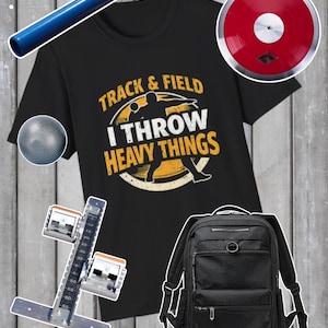 以下が含まれることがあります： 「Track & Field I Throw Heavy Things」のグラフィックが入った黒いTシャツ。青いバトン、赤い円盤、砲丸投げ、スターティングブロック、黒いバックパックが含まれています。陸上競技愛好家向けです。