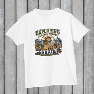 May include: A white short-sleeve t-shirt featuring a graphic print of a young explorer holding a map in front of mountains and a compass. The text reads Exploring My Way Through 6th Grade A Adventure Awaits, with illustrations of binoculars, pencils, and hiking boots.