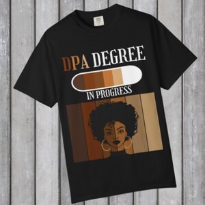 May include: Black t-shirt with the text "DPA DEGREE IN PROGRESS" in white and brown. The shirt features a graphic of a woman's face with large hoop earrings and a gradient of skin tones.