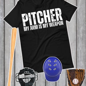May include: Black t-shirt with the text "PITCHER MY ARM IS MY WEAPON" in white. A baseball bat, catcher's mask, blue helmet, and baseball glove with a baseball are also displayed. The items are arranged on a wooden background.