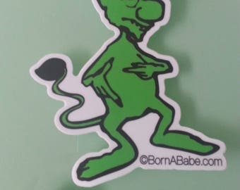 Puede incluir: Una pegatina de duende verde de dibujos animados con un borde blanco. El duende tiene orejas grandes, una nariz larga y una cola curvada. La pegatina incluye el texto "©BornABabe.com". Una moneda está al lado de la pegatina para la comparación de tamaño.