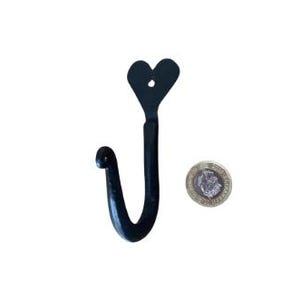 May include: Black metal wall hook with a heart-shaped top. The hook is curved and has a rounded end. A coin is next to the hook for size reference. The hook is likely for hanging items like coats or bags.