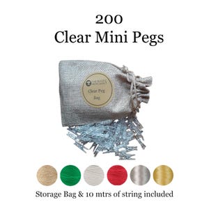 May include: A set of 200 clear mini pegs in a beige drawstring storage bag with a circular label reading The Rustic Merchant Clear Peg Bag. Below the bag are six spools of string in beige, green, white, red, grey, and gold, with 10 metres included.