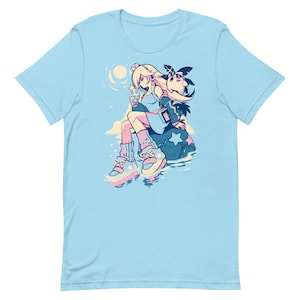 Op de afbeelding: Een lichtblauw t-shirt met een afbeelding van een cartoonfiguur met roze haar en een blauwe jurk, zittend op een rots met een ster erop. Het personage houdt een klein blauw voorwerp vast en heeft een roze en blauwe vogel op zijn schouder.