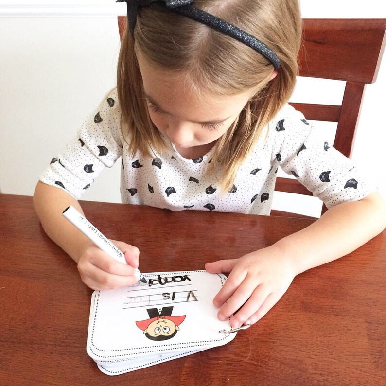May include: A child is writing the word "vampire" on a white card with a cartoon vampire illustration. The card says "V is for..." and has a ring binder.