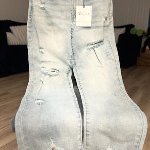 May include: Light blue denim jeans with distressed details, including rips and a frayed hem. A tag from "21 Special A" hangs from the waistband. The jeans are displayed on a chair.