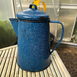 Peut inclure: Une cafetière en émail bleu avec une finition mouchetée et une poignée. La cafetière est posée sur une surface en bambou brun clair.