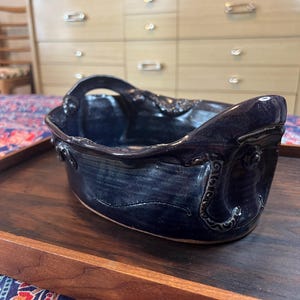 Puede incluir: Un plato de cerámica azul oscuro con asa. El plato tiene forma rectangular con detalles ornamentados en los bordes. Se encuentra sobre una bandeja de madera oscura.