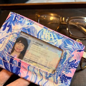 May include: A rectangular fabric ID holder featuring a vibrant blue, pink, and white abstract floral pattern. The front includes a clear plastic window displaying an Oregon driver license, with a gold-toned metal ring attached to the side.