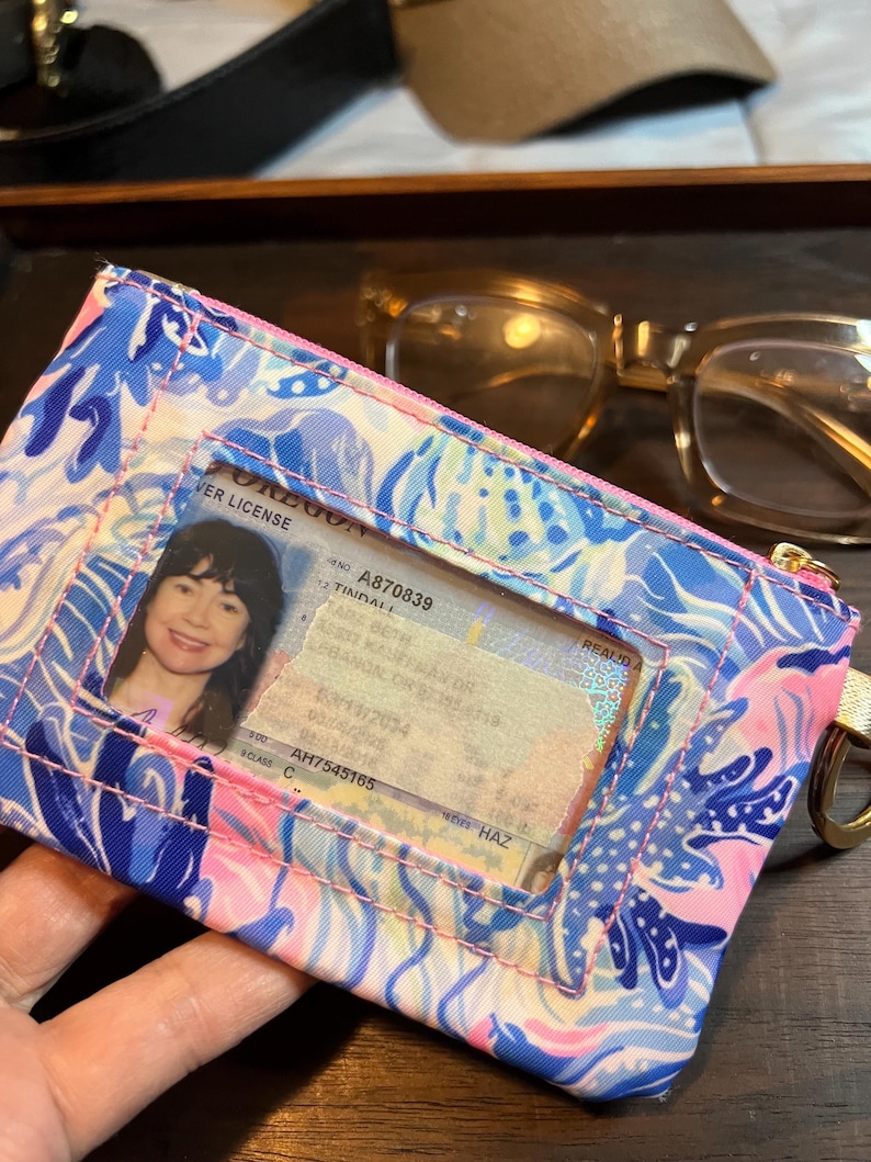 May include: A rectangular fabric ID holder featuring a vibrant blue, pink, and white abstract floral pattern. The front includes a clear plastic window displaying an Oregon driver license, with a gold-toned metal ring attached to the side.