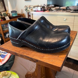May include: A pair of black leather clogs with a rounded toe, a low heel, and visible white stitching along the base of the upper. The shoes feature a smooth finish and a sturdy construction with a dark, solid sole.
