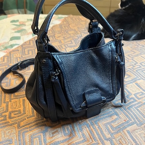 May include: A dark navy blue handbag with a pebbled texture, featuring a structured top handle, a detachable shoulder strap, and a front flap pocket secured with a metal buckle. The bag has dual side zippers with long leather pulls and dark hardware.