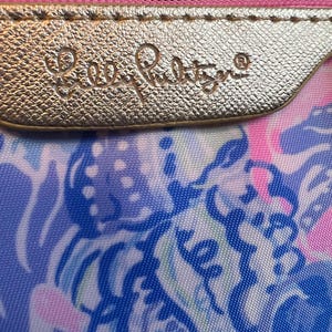 May include: A zippered pouch featuring a gold-toned metallic strip embossed with the text Lilly Pulitzer. The pouch displays a vibrant, multi-colored abstract pattern in shades of blue, pink, and white, with a pink zipper lining the top edge.