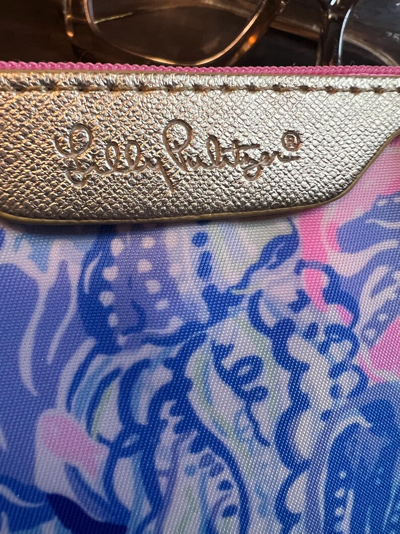 May include: A zippered pouch featuring a gold-toned metallic strip embossed with the text Lilly Pulitzer. The pouch displays a vibrant, multi-colored abstract pattern in shades of blue, pink, and white, with a pink zipper lining the top edge.