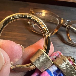 May include: A gold-toned metal keyring featuring the engraved text LILLY PULITZER. The circular ring is attached to a fabric accessory with a pink, blue, and white patterned design, secured by a metallic gold-colored connector piece.